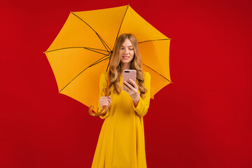 happy young woman in yellow dress with umbrella using mobile phone on red background