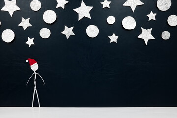 A solo stick man figure in santa hat alone in a black background with silver Christmas ornaments.
