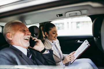 Good looking senior business man and his young woman colleague or coworker sitting on backseat in luxury car. They talking, smiling and using laptop and smart phones. 