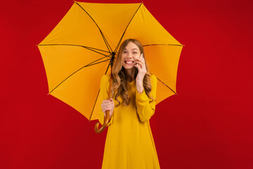 Beautiful happy young woman in a yellow dress with an umbrella, talking on a mobile phone on a red background