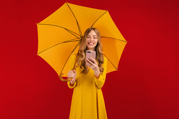 Beautiful happy young woman in yellow dress with umbrella using mobile phone on red background