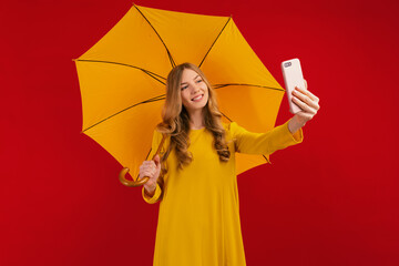 Funny cute young woman in a yellow dress with an umbrella, taking a selfie on a mobile phone on a red background
