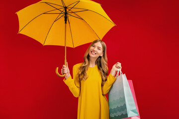 Happy beautiful young woman with a yellow umbrella and shopping bags on a red background, shopping concept, autumn