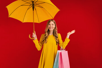 Excited shaken young woman, with a yellow umbrella and shopping bags on a red background, shopping concept, autumn
