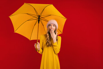 Shocked beautiful young woman with an umbrella in a warm hat on her head, on a red background