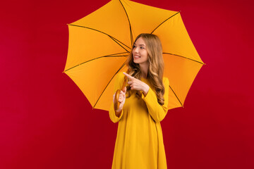 happy young woman with a yellow umbrella pointing at a copy of the space on an empty red background