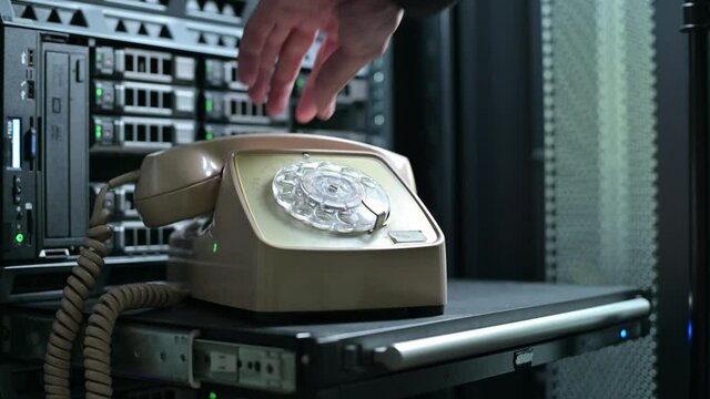 A man puts an old rotary telephone on a shelf against the backdrop of a modern server cabinet and starts dialing a phone number. Clash of eras, progress and technology
