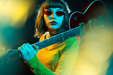 Stylish young hipster woman with curly hair holding red guitar on shoulder in neon lights. 90s...