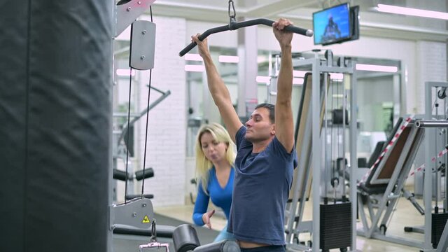 coach helps the client to control the correctness of the exercise in the gym. Skinny guy under the supervision of an athletic young woman