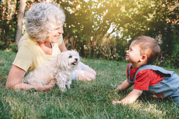 Grandmother lying on grass with grandson boy at home backyard. Bonding of relatives and generation communication. Old woman with baby having fun spending time together outdoors. - 374217827