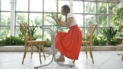 young blonde woman in long red skirt with glasses surfs internet with contemporary tablet sitting at table in cafe with pot plants