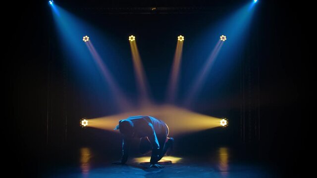 Young sporty guy dancing break dance on the background of studio lighting. Silhouette dancer man in a tracksuit performs complex movements. Street dancing as a lifestyle. Slow motion.