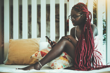 An adorable young black woman in eyeglasses, nightie, and with long braids of a dark-red color is sitting on her bed in the morning at home and reading newsfeed on the screen of her smartphone - Powered by Adobe