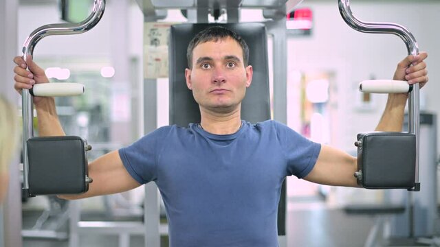 A thin guy does an exercise on an exercise machine under the supervision of a gym trainer, the camera moves back and forth, front view