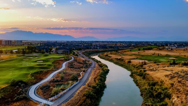 Aerial hyperlapse over a river with the rising sun and an idyllic community with a golf course, commuter train and a city in the valley