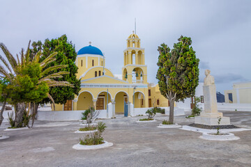 Fototapeta na wymiar Classical Greek Orthodox church in Oía, Santorini island. Beautiful yellow church with blue dome in a square with green trees in cloudy day.