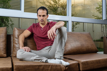 Sad and downcast Brazilian mature man (44 years old), sitting on the brown sofa, with a large window pane behind.