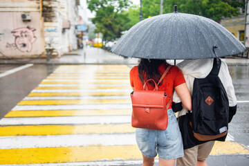 Young couple in love under one umbrella crossing the pedestrian in bad rainy weather. Rainy day. Umbrella with raindrops. City scenes in the rain.