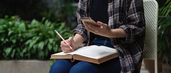 Female student reading book and using smartphone in garden