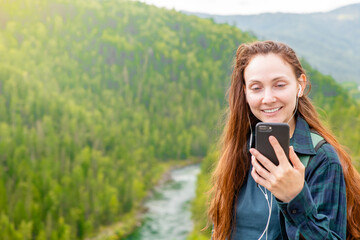 Smiling woman wearing headphones holds smartphone at mountains and listening to audio guide