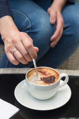 hand with a ring spooning a coffee with foam inside a small white cup that has a plate and napkin, on a smooth black surface, in the background you can see the body of a woman
