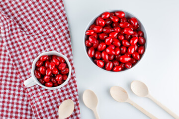 Cornel berries in  bowl and cup with wooden spoons flat lay on picnic cloth and white background