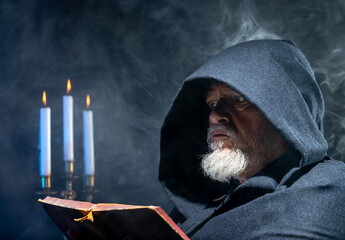 Portrait of an elderly man with a beard. He is wearing a hooded cloak and is reading a book. A...