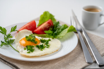 Healthy and hearty breakfast. Close-up of a fried eggs, tomato, herbs and espresso coffee. Served on a white table with a linen napkin and cutlery.