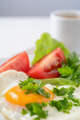 Healthy and hearty breakfast. Close-up of a fried eggs, tomato, herbs and espresso coffee. Served on a white table with a linen napkin and cutlery.