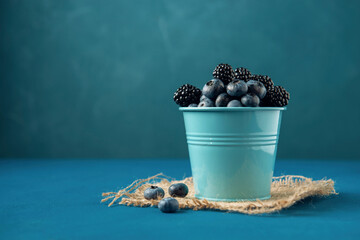Fototapeta na wymiar Small bucket with blueberry and blackberry on burlap napkin and blue background.