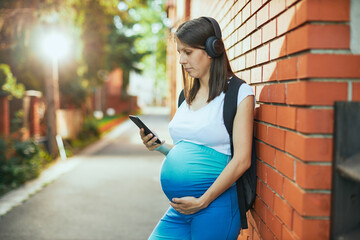 Fototapeta premium Pregnant woman standing outdoors, using smart phone and listening music. She is in sportswear.