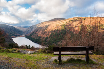 View over Upper Lake in Glendalough Scenic Park, a lot of hills, sun and clouds, bench on the front. Concepts: landscape, outdoor, tourism, travel