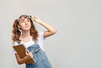 Curious student portrait. Course advertising. Smart girl with notebook taking off eyeglasses learning new information looking up at empty space isolated on neutral background. Class presentation.