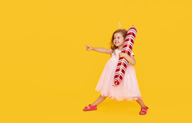 A cute little girl child in a fancy dress and a Santa hat holds an inflatable shape of a candy cane on a yellow background. 2021 new year holidays concept. points to the left