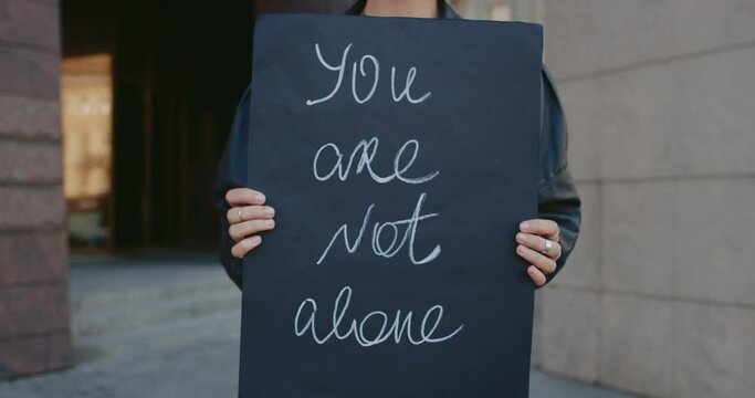 Female hands holding carton placard with you are not alone writing on it at city street. Concept of social problem and support