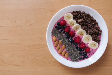 Smoothie bowl with fresh raspberry, blueberry, chia seeds, almonds, banana, and chocolate flakes. Morning breakfast on wooden table. Top view