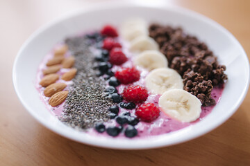 Smoothie bowl with fresh raspberry, blueberry, chia seeds, almonds, banana, and chocolate flakes. Morning breakfast on wooden table. Close up
