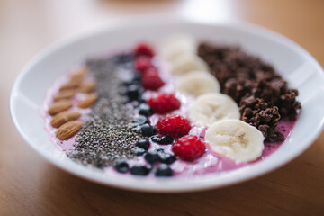 Smoothie bowl with fresh raspberry, blueberry, chia seeds, almonds, banana, and chocolate flakes. Morning breakfast on wooden table. Close up