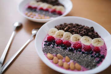 Smoothie bowl with fresh raspberry, blueberry, chia seeds, almonds, banana, and chocolate flakes. Morning breakfast on wooden table. Two ptales fith smoothie bows. Vegan food