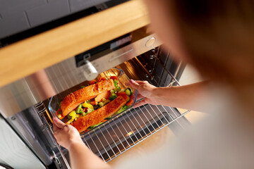 culinary, food and people concept - woman cooking salmon fish with vegetables in baking dish in oven at home kitchen
