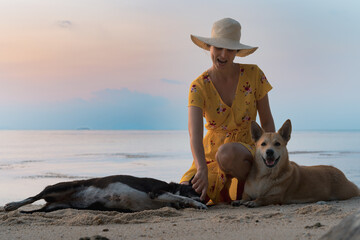 Young caucasian woman in hat and yellow dress sits on the sandy beach and playing with dogs at sunset