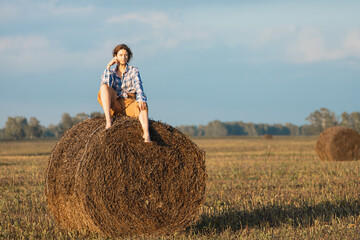 Naklejka na ściany i meble Beautiful modern young woman wearing in beige trousers and a plaid shirt posing against a haystack in field. Trendy teenage girl in autumn outdoors.