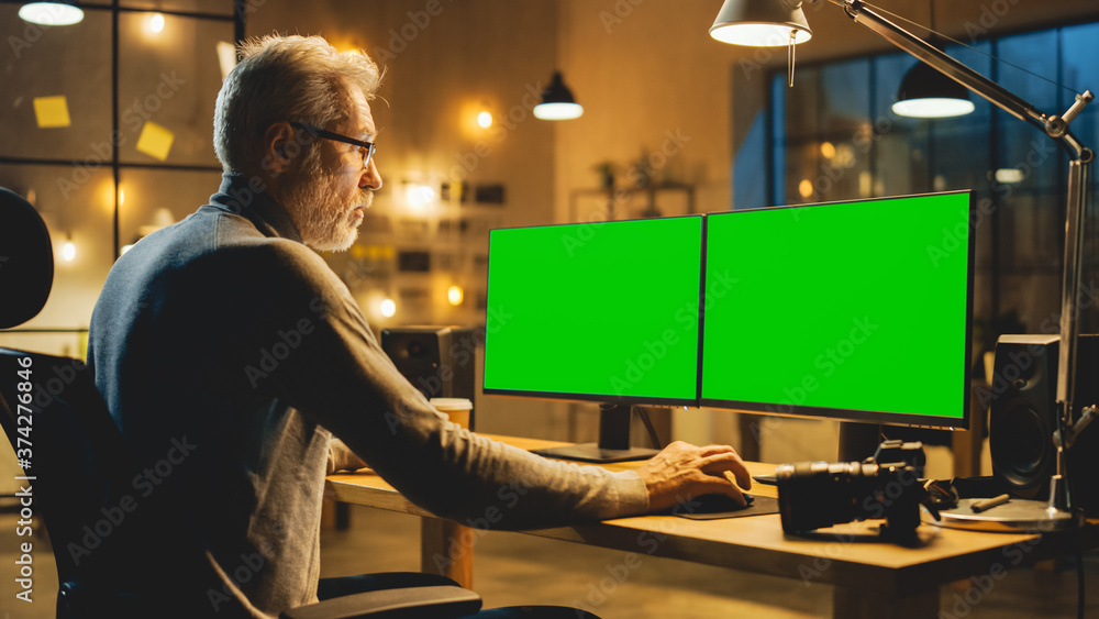Poster Creative Middle Aged Designer Sitting at His Desk Uses Desktop Computer with Two Green Mock-up Screens. Professional Office Employee Working Late in the Evening in His Studio 