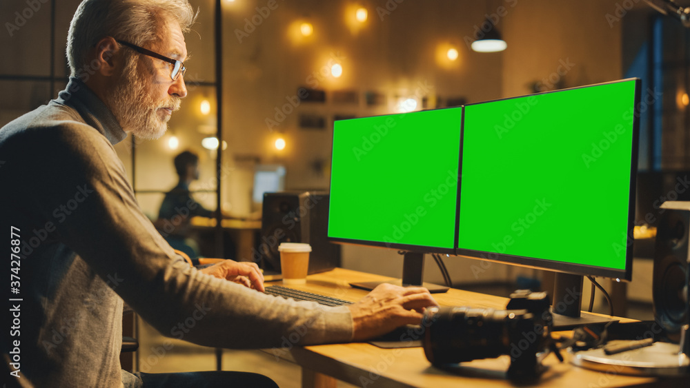 Poster creative middle aged designer sitting at his desk uses desktop computer with two green mock-up scree
