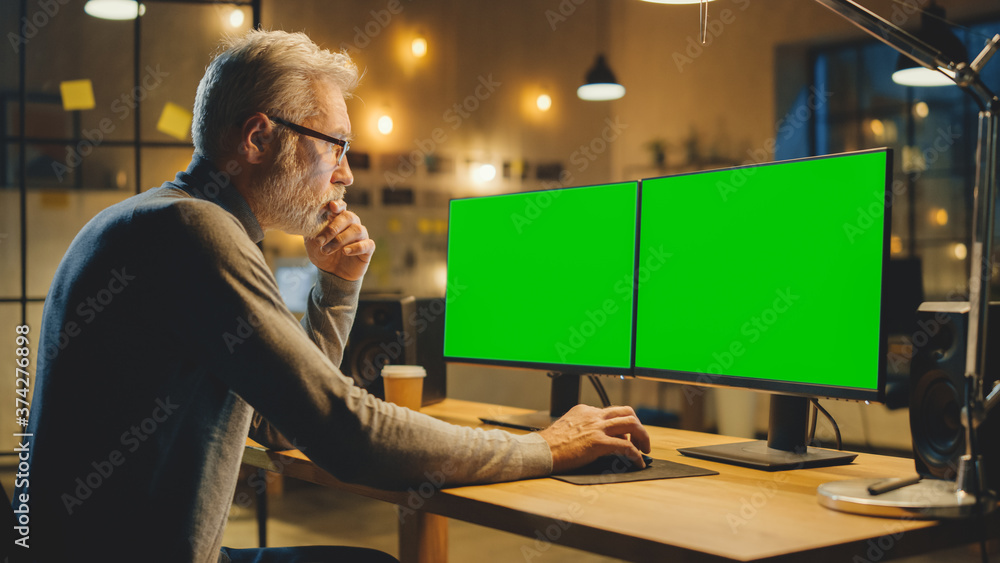 Poster Creative Middle Aged Designer Sitting at His Desk Uses Desktop Computer with Two Green Mock-up Screens. Professional Office Employee Working Late in the Evening in His Studio 