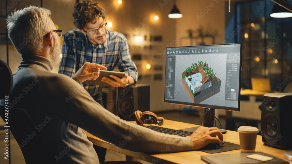 Poster In the Evening Creative Middle Aged Video Game Developer Works on a Desktop Computer with Screen 3D Videogame Level Design, Has Conversation with Younger Specialist who Uses Tablet Computer