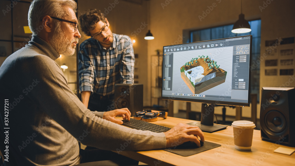 Poster In the Evening Creative Middle Aged Video Game Developer Works on a Desktop Computer with Screen 3D Videogame Level Design, Has Conversation with Younger Specialist who Uses Tablet Computer