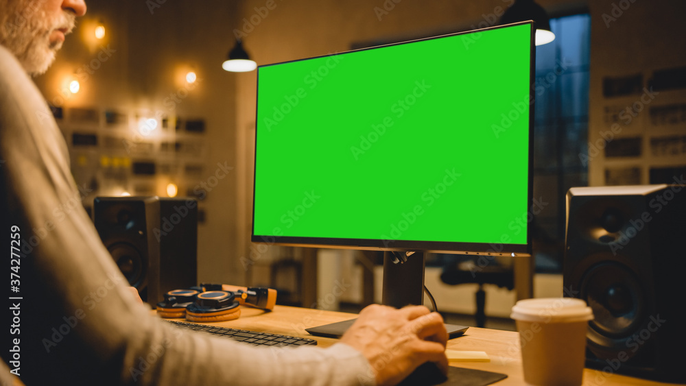 Poster Creative Middle Aged Designer Sitting at His Desk Uses Desktop Computer with Two Green Mock-up Screens. Professional Office Employee Working Late in the Evening in His Studio 