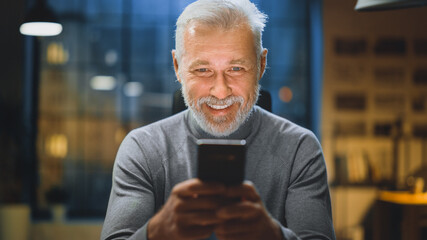 Portrait of the Handsome and Successful Middle Aged Bearded Businessman Uses Smartphone while Sitting at His Desk, Laughs and Smiles. Working from Cozy Home Office / Studio 