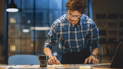 Young Creative Designer Working at His Desk, Concentrated on the Paperwork, Choosing Right Design, Concept and Sketches. In the Background Evening Office Studio with Window